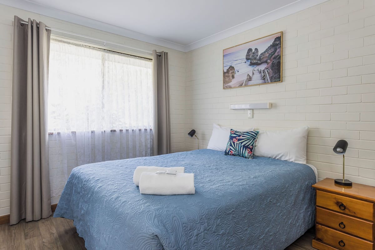 A bedroom with a large bed, a nightstand, a lamp, a painting on the wall, and a window with curtains.