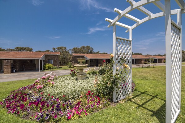 Un espace jardin agrémenté d’une pergola blanche, de parterres de fleurs et d’un bâtiment doté d’un toit rouge.