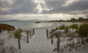 Une plage de sable avec une clôture en bois au premier plan et une vue sur l’océan et les collines lointaines.