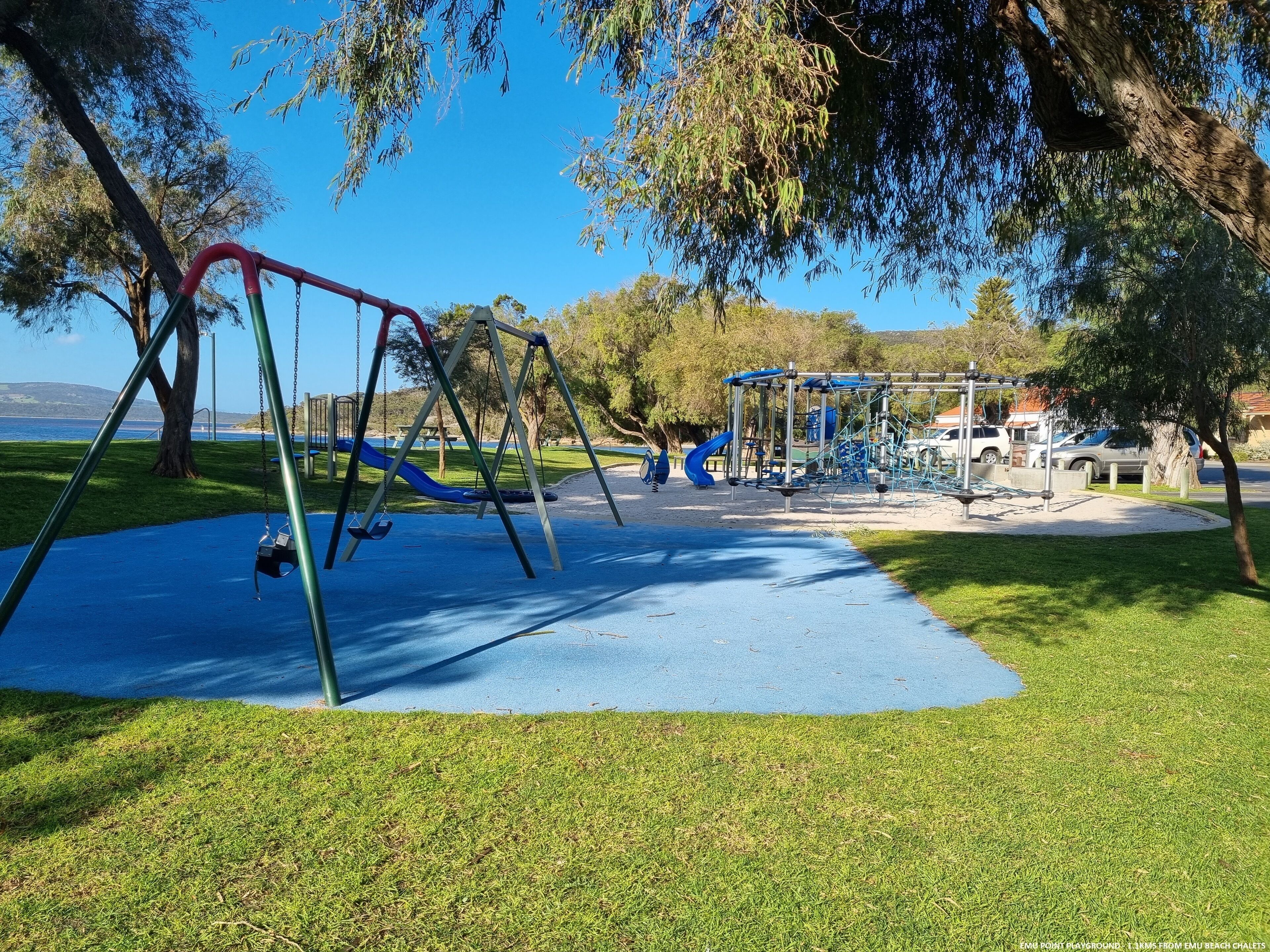 A playground with swings and slides, surrounded by grass and trees, with a clear view of the sky.