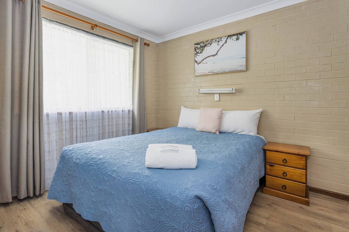 A bedroom with a bed, a nightstand, a wall-mounted light, and a window with curtains.