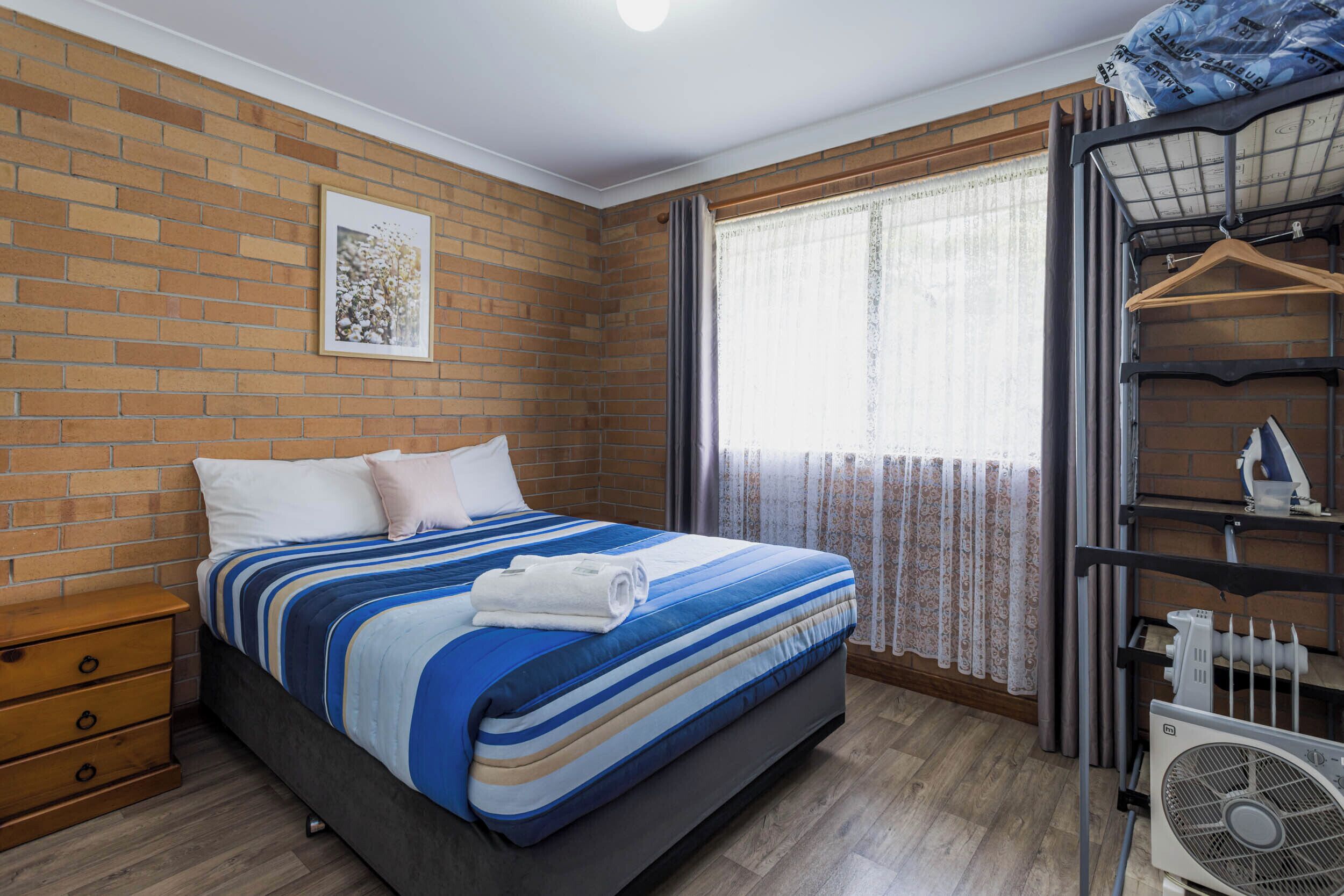 A bedroom with a brick wall, a bed with a striped bedspread, a nightstand, a window with curtains, and a radiator.