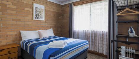 A bedroom with a brick wall, a bed with a striped bedspread, a nightstand, a window with curtains, and a radiator.