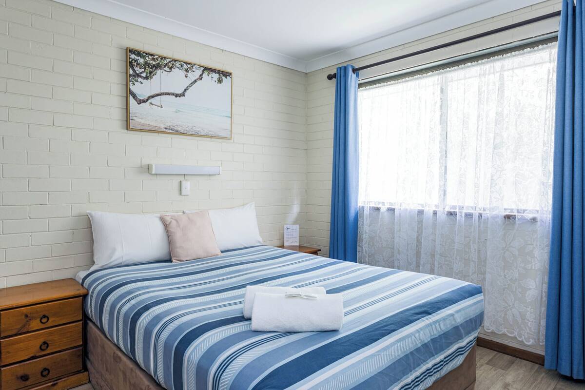 A bedroom with a bed, a nightstand, a wooden dresser, a wall-mounted picture, and a window with curtains.