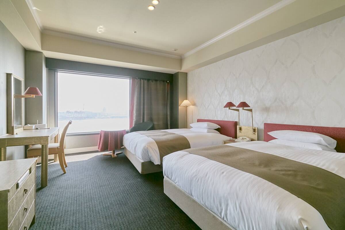 Habitación de hotel con dos camas, un escritorio, una silla y un amplio ventanal con vistas a la ciudad.