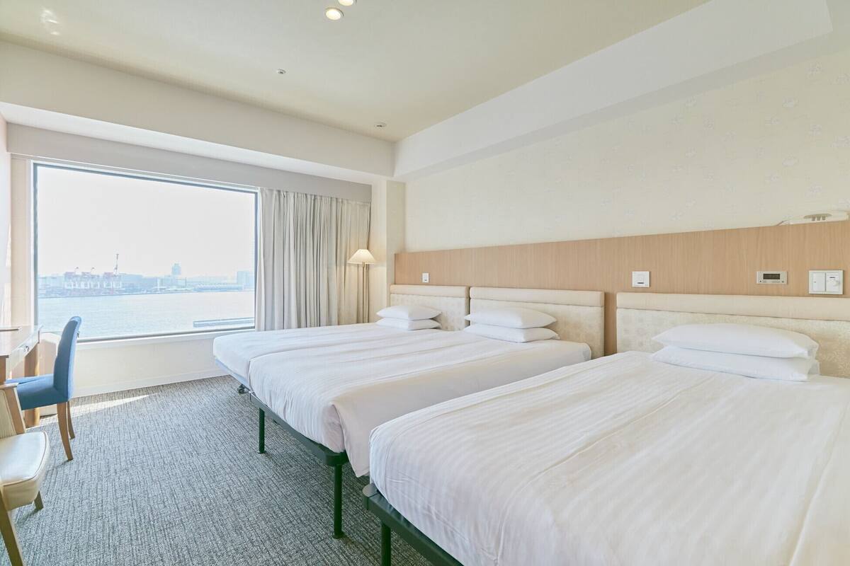 Habitación de hotel con dos camas, un escritorio y vistas a un paisaje urbano.