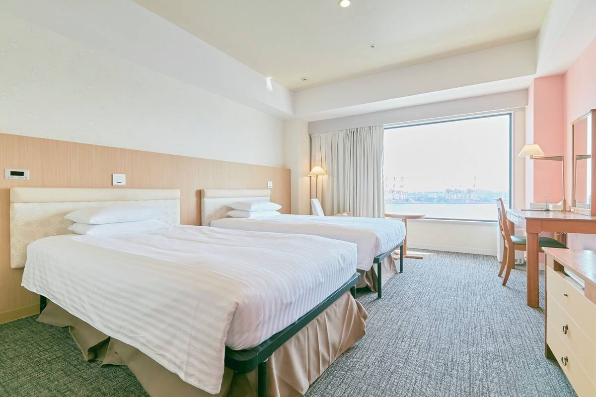 Habitación de hotel con dos camas, un escritorio, una silla y un amplio ventanal con vistas a la ciudad.