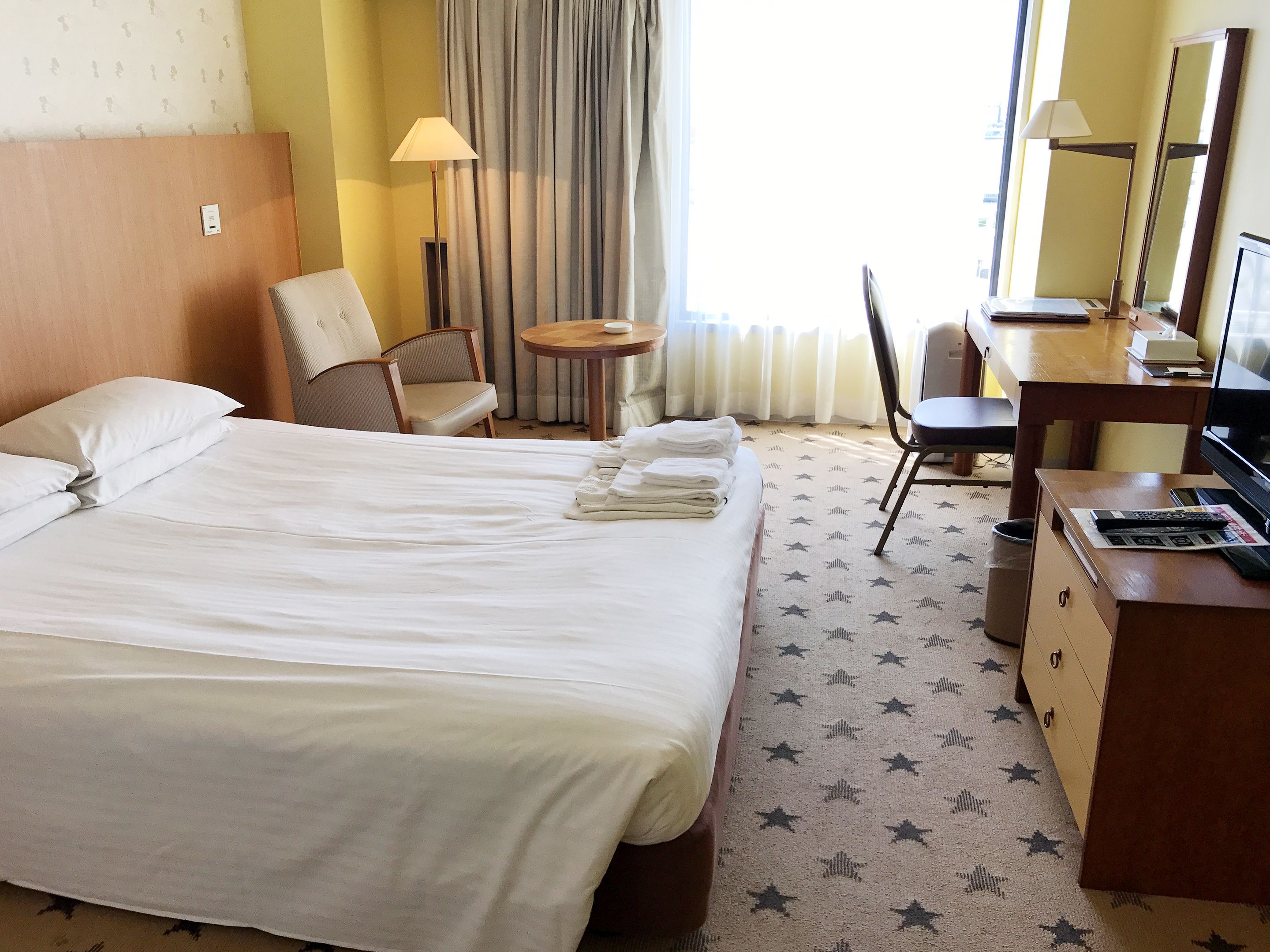 A hotel room with a large bed, a desk, a chair, a small table, and a lamp.