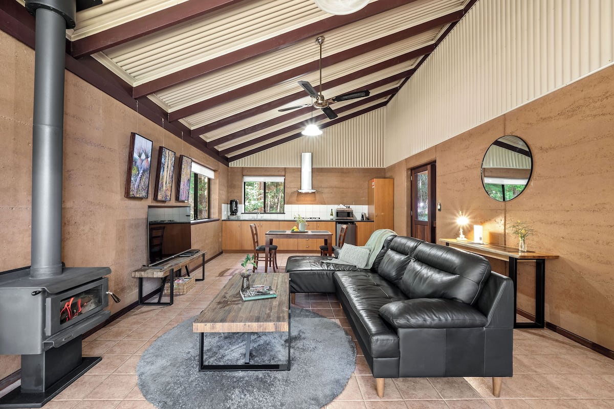 A spacious living room with a fireplace, a black leather sofa, a wooden coffee table, and a kitchen in the background.
