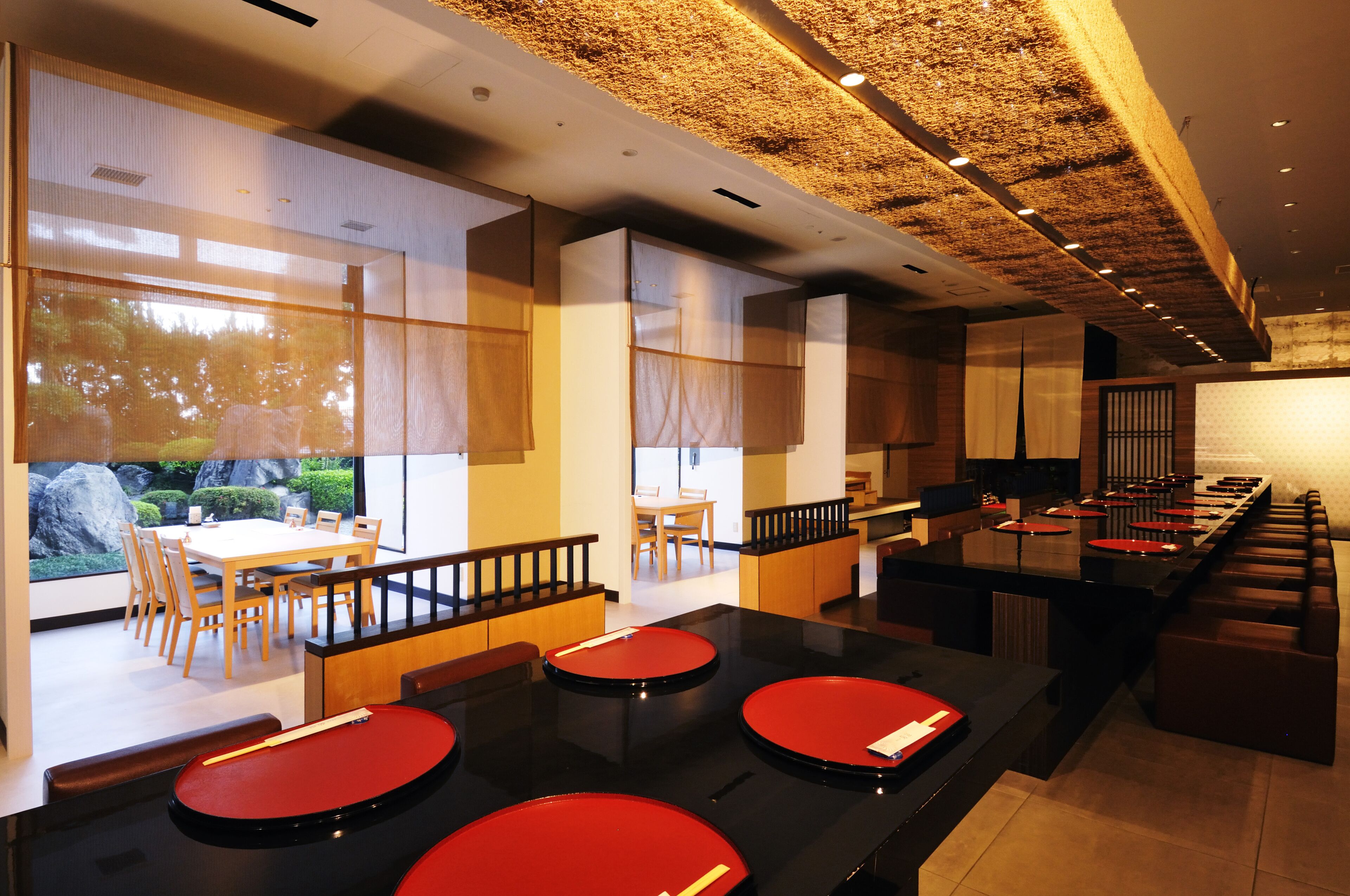 Un restaurant moderne avec un long comptoir, des tabourets rouges et une vue sur un jardin grâce à de grandes fenêtres.