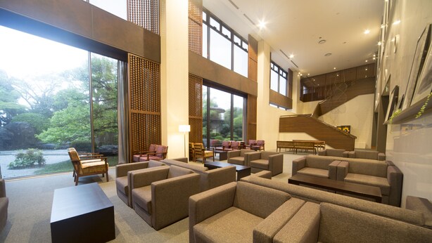 A spacious lobby with multiple seating areas, large windows offering views of greenery, and a staircase leading to an upper level.