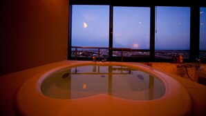 A modern bathroom with a large bathtub, a view of the cityscape through large windows, and a balcony railing.