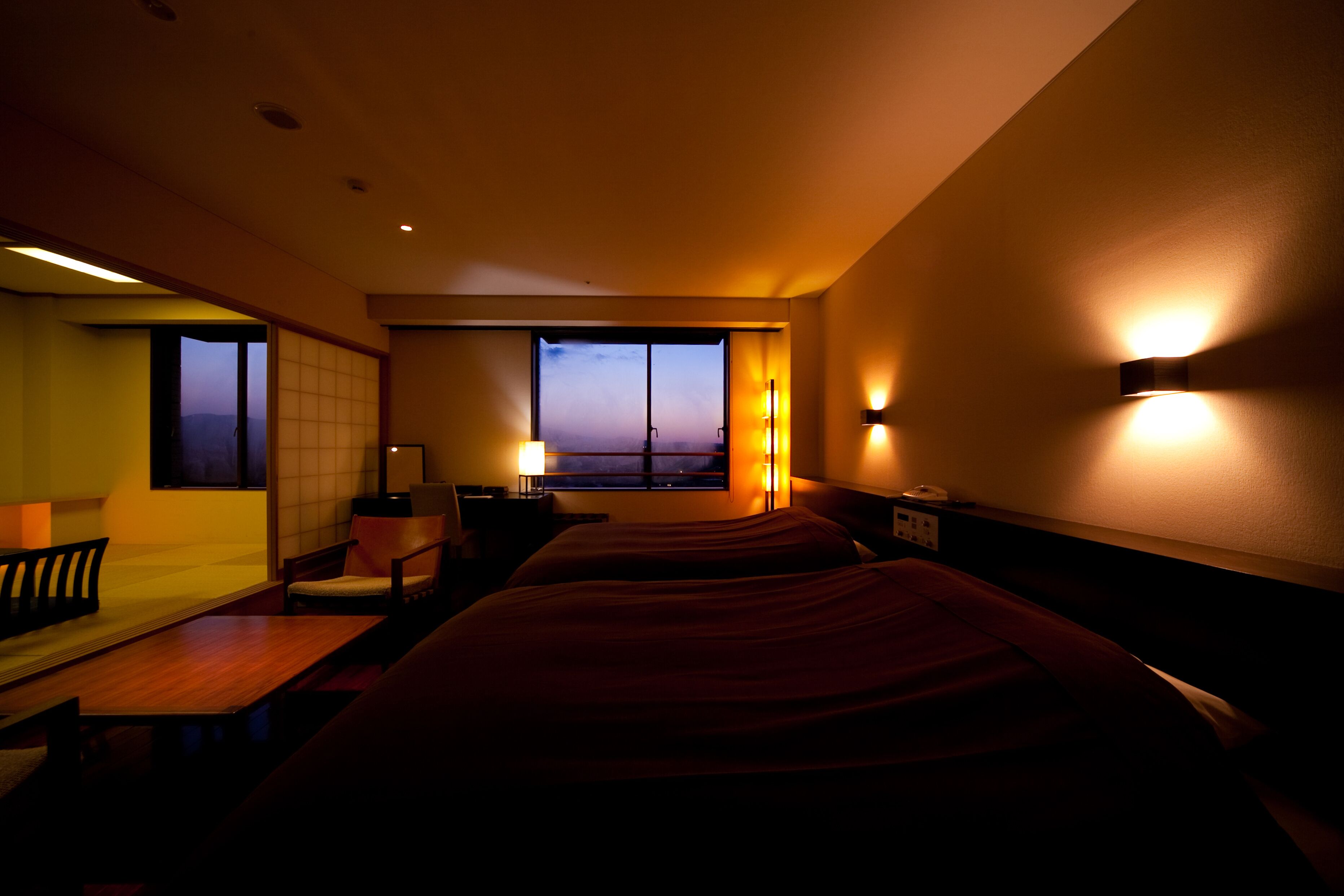 A hotel room with a large bed, a desk, a chair, and a view of the sunset through the window.