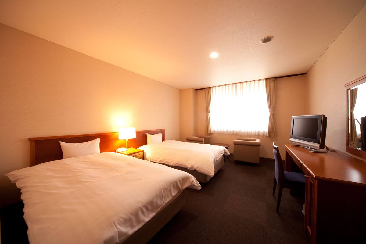 Una habitación de hotel con dos camas, un escritorio con televisor y una ventana con cortinas.