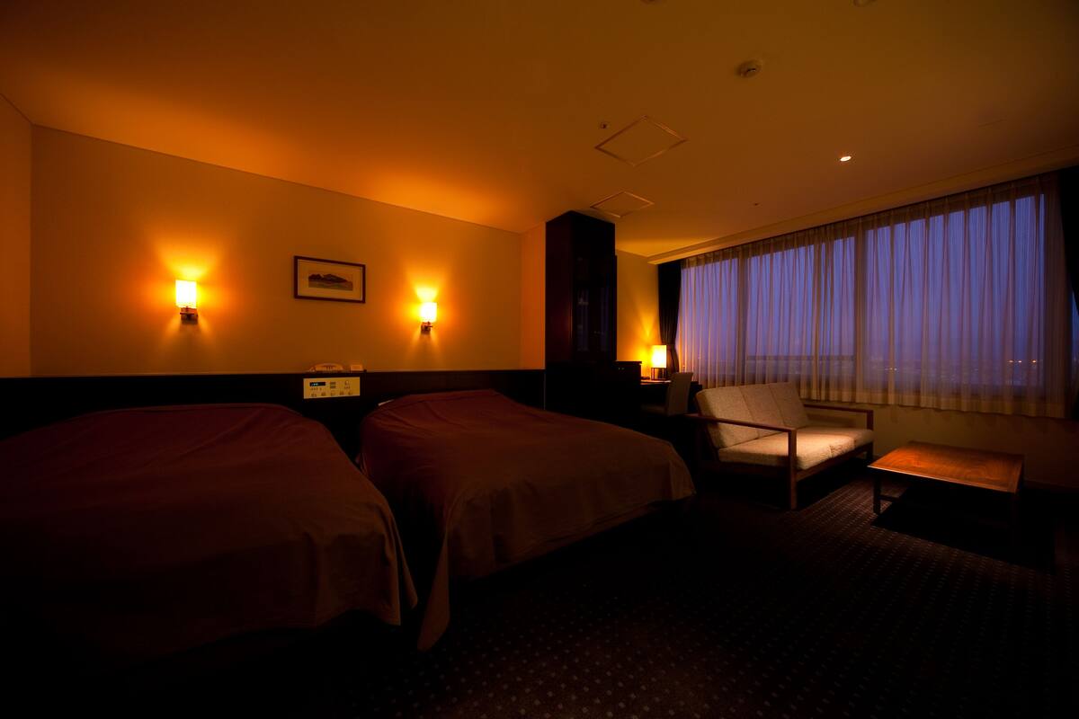 Habitación de hotel con dos camas, una silla, una mesita y una ventana con cortinas.