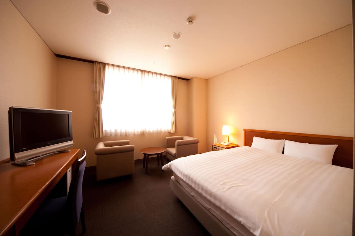 Habitación de hotel con cama, escritorio con televisor, dos sillones, una mesita y una mesita de noche con lámpara.