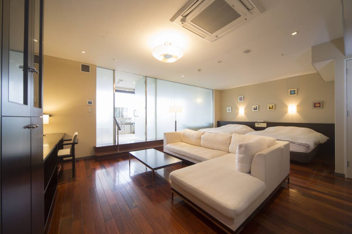 Habitación de hotel moderna con suelo de madera, sofá, cama, escritorio y una silla. Cuenta con un ventanal grande y una puerta que da a un balcón.
