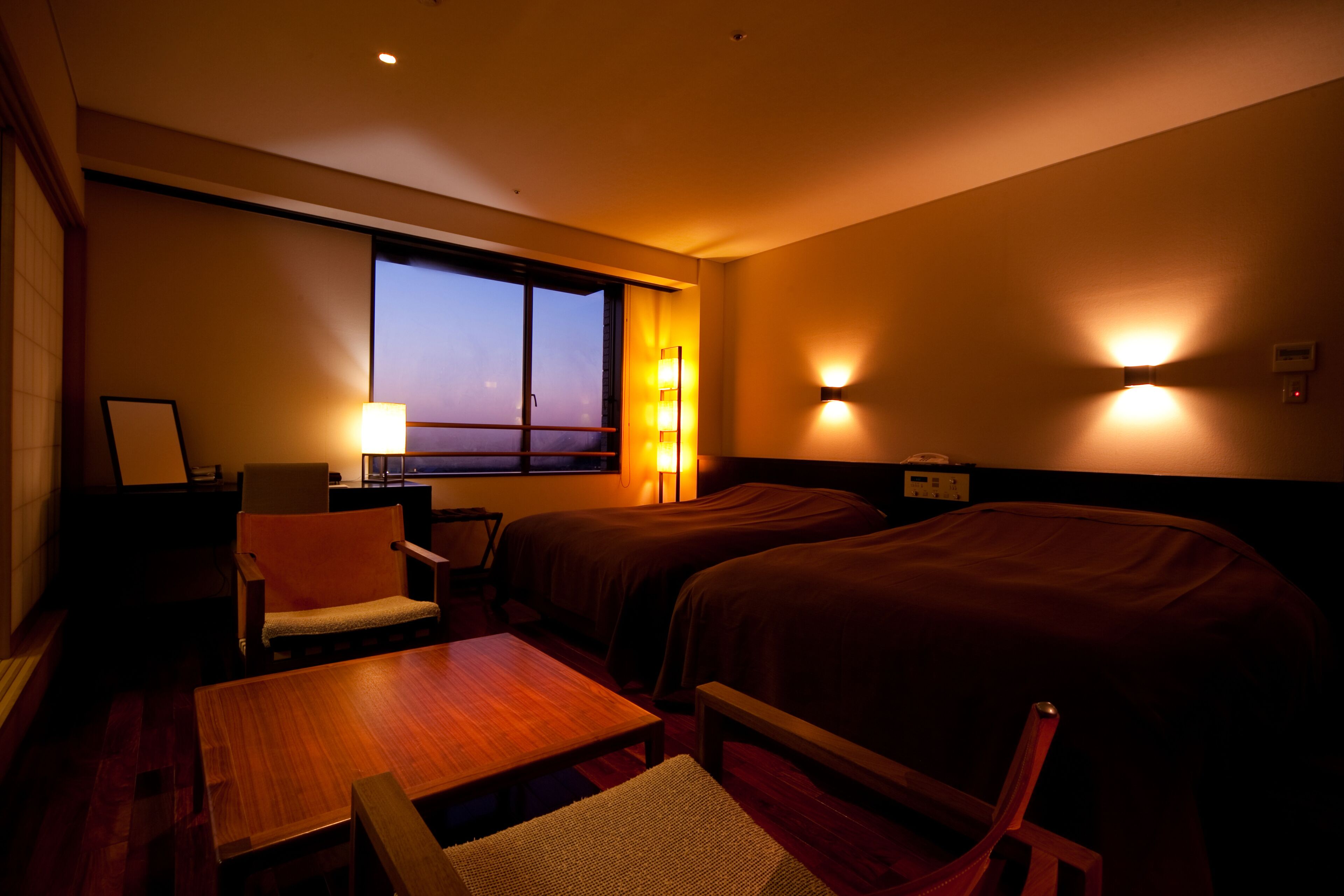 Ein Hotelzimmer mit zwei Betten, einem Holztisch, einem Stuhl, einem Schreibtisch mit Lampe und Meerblick durch ein Fenster.