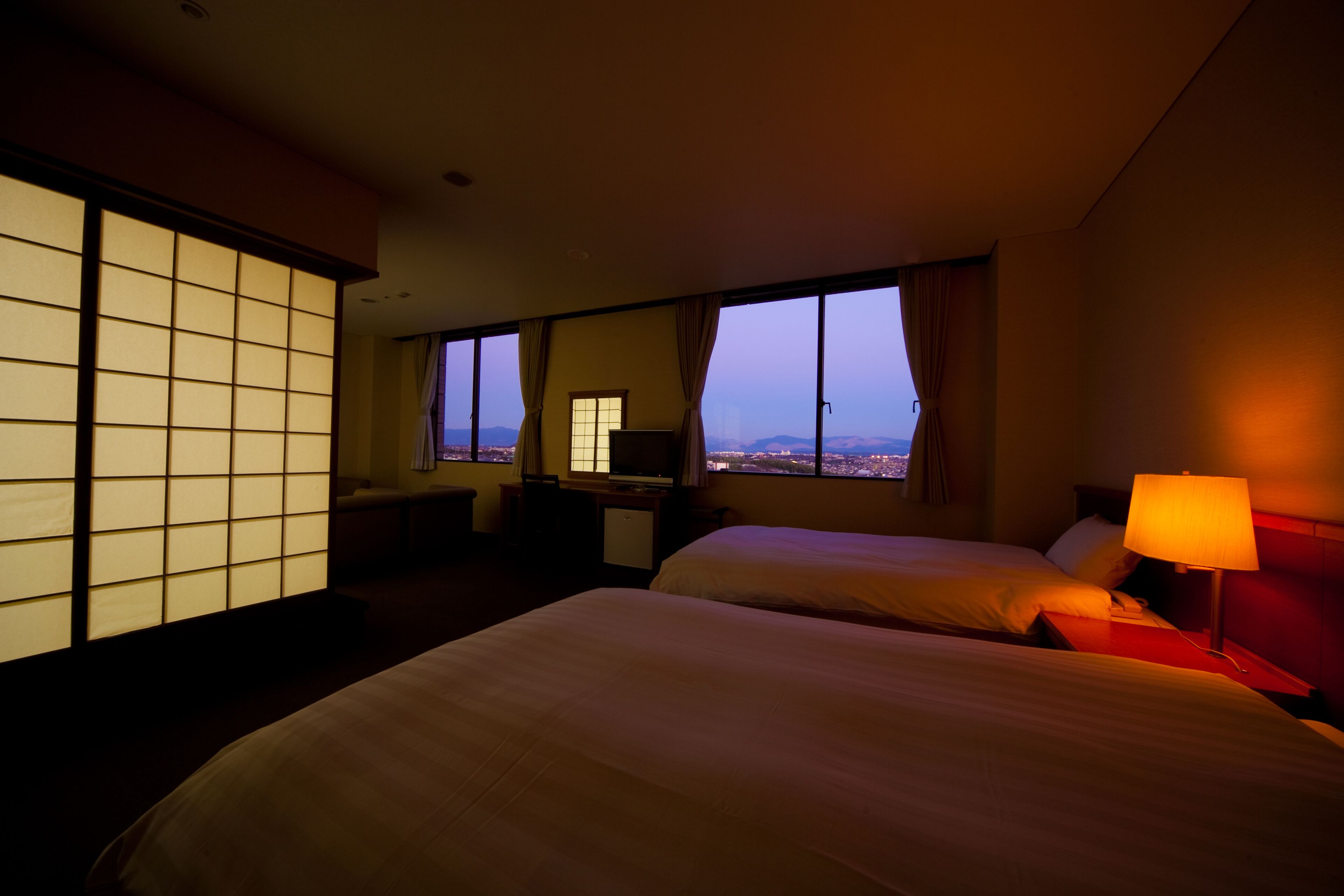 A hotel room with two beds, a desk, a TV, and a view of the cityscape.