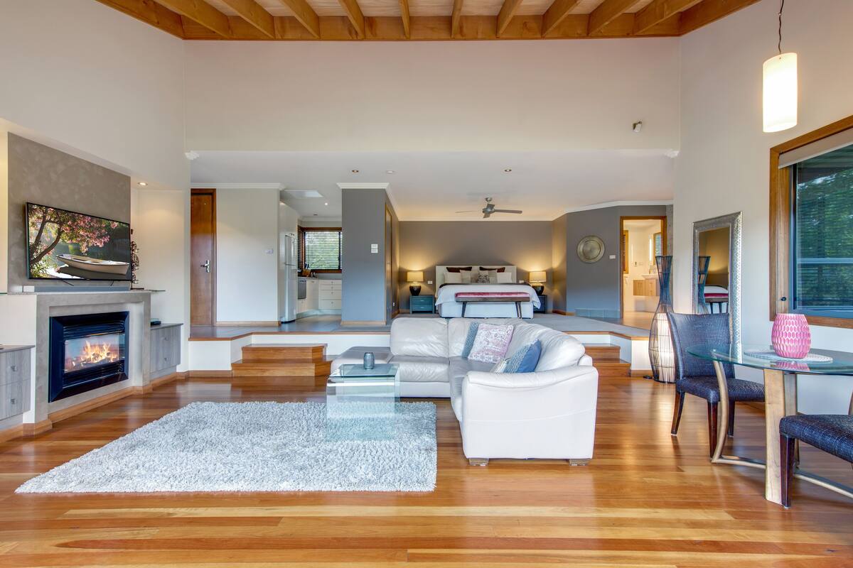 A spacious living room with a fireplace, white sofas, a glass coffee table, and a dining area with a glass table and chairs.