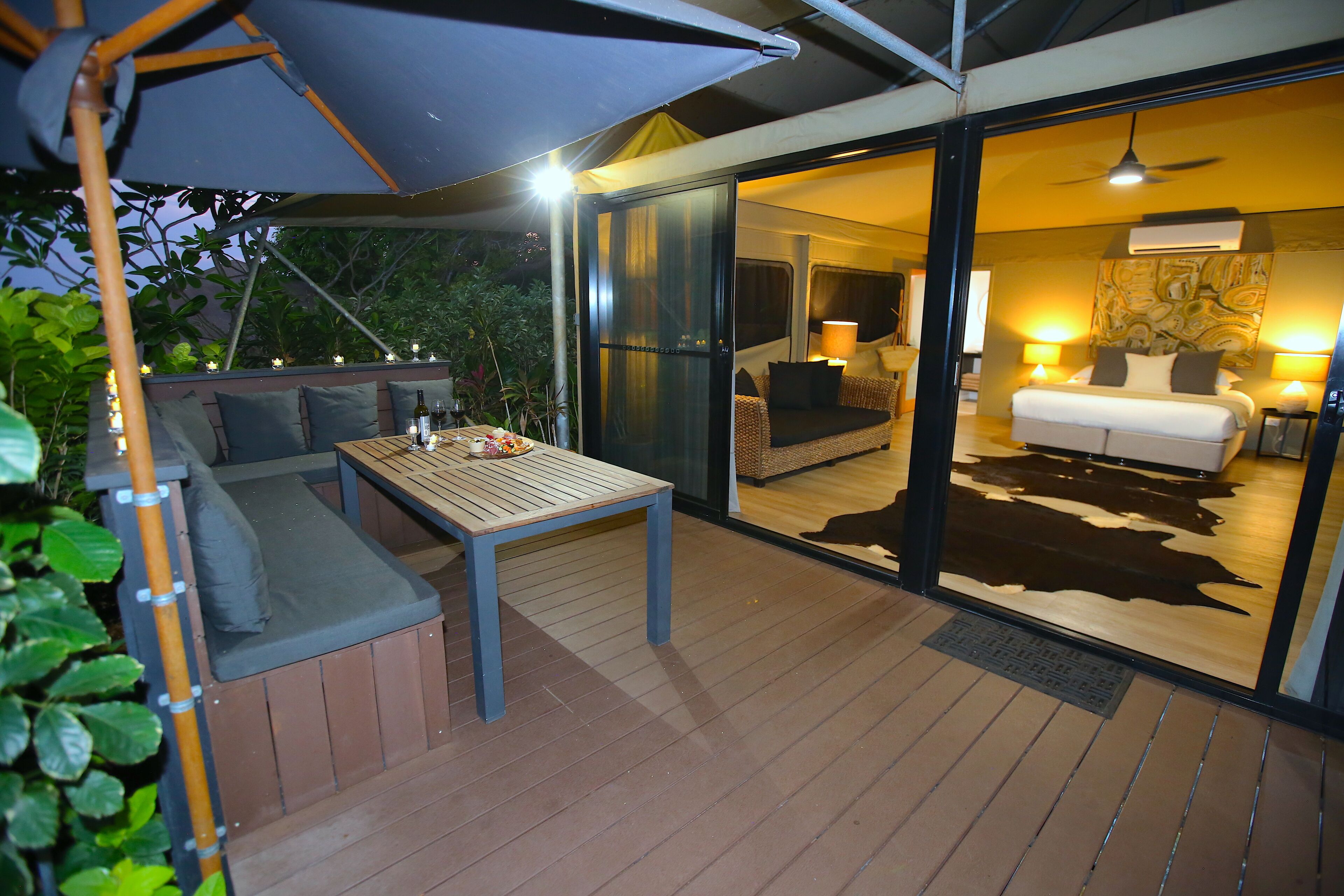 An outdoor seating area with a table and cushions, a glass door leading to a bedroom with a bed and two bedside tables, and a ceiling fan.
