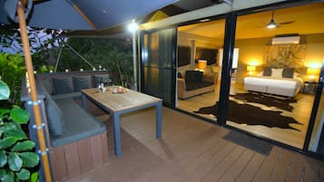 Un'area salotto esterna con tavolo e cuscini, una porta vetrata che dà su una camera da letto con un letto e due comodini, e una ventola a soffitto.