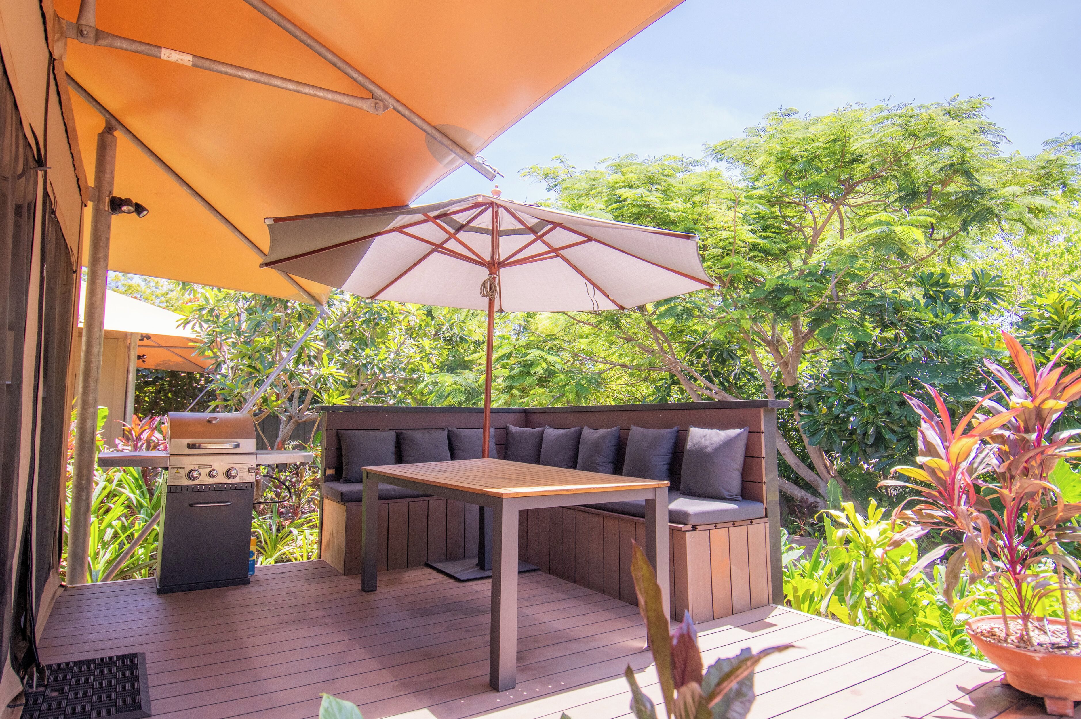 Eine Terrasse im Freien mit Holztisch, Grill und einer Sitzgruppe mit Polstern unter einem Sonnenschirm.