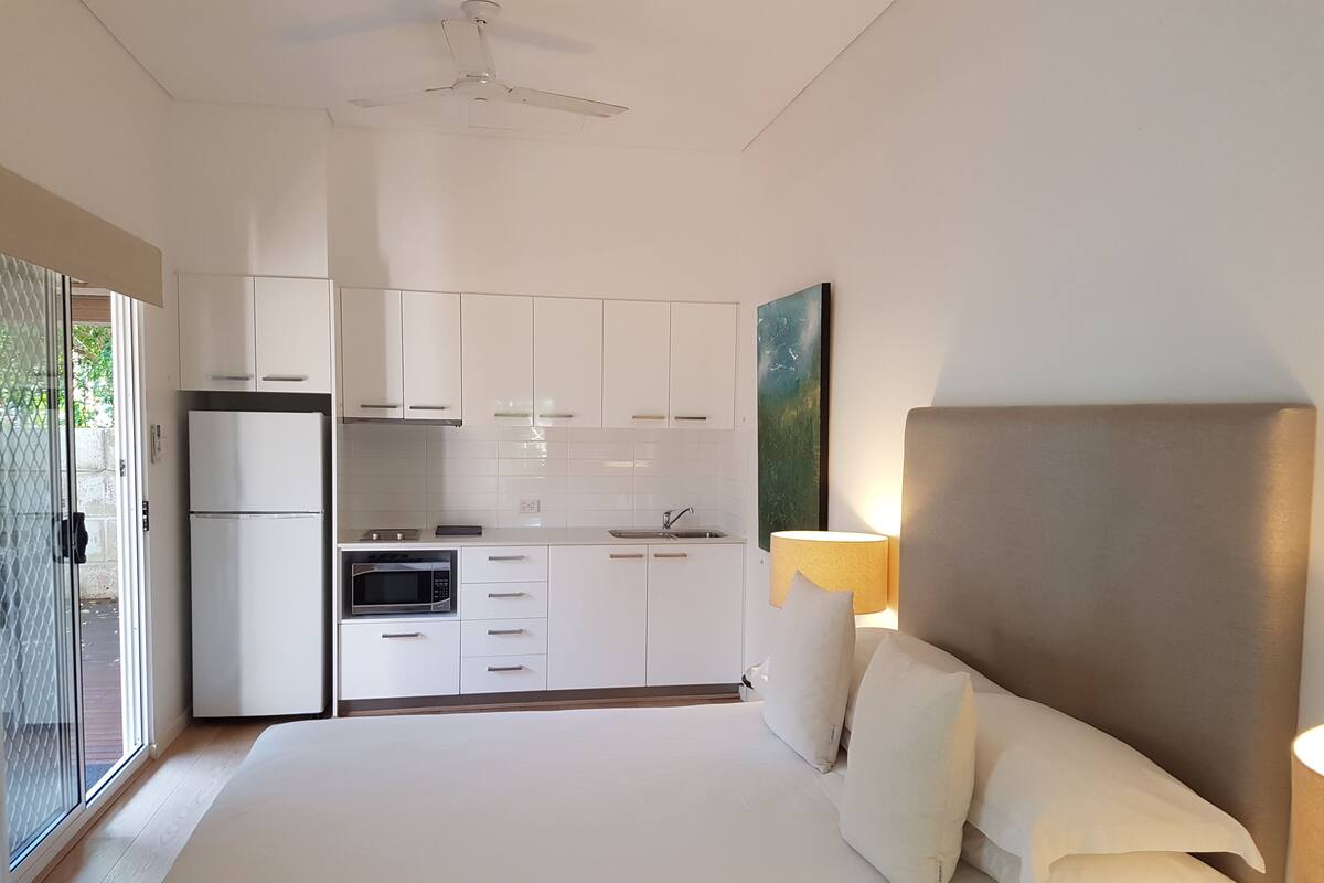 A modern bedroom with a large bed, a bedside lamp, and a view of a kitchen with white cabinets and a refrigerator.