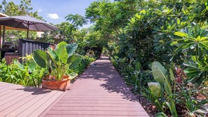 A wooden pathway flanked by lush tropical plants, leading to a shaded area with a large umbrella. - The Billi Resort (Cable Beach)