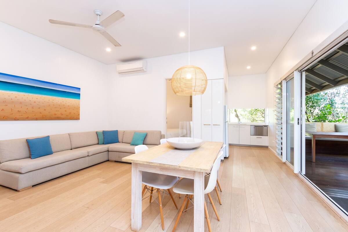 A modern living room with a wooden dining table, white chairs, a beige sofa with blue cushions, a ceiling fan, and a large window with a view of greenery.