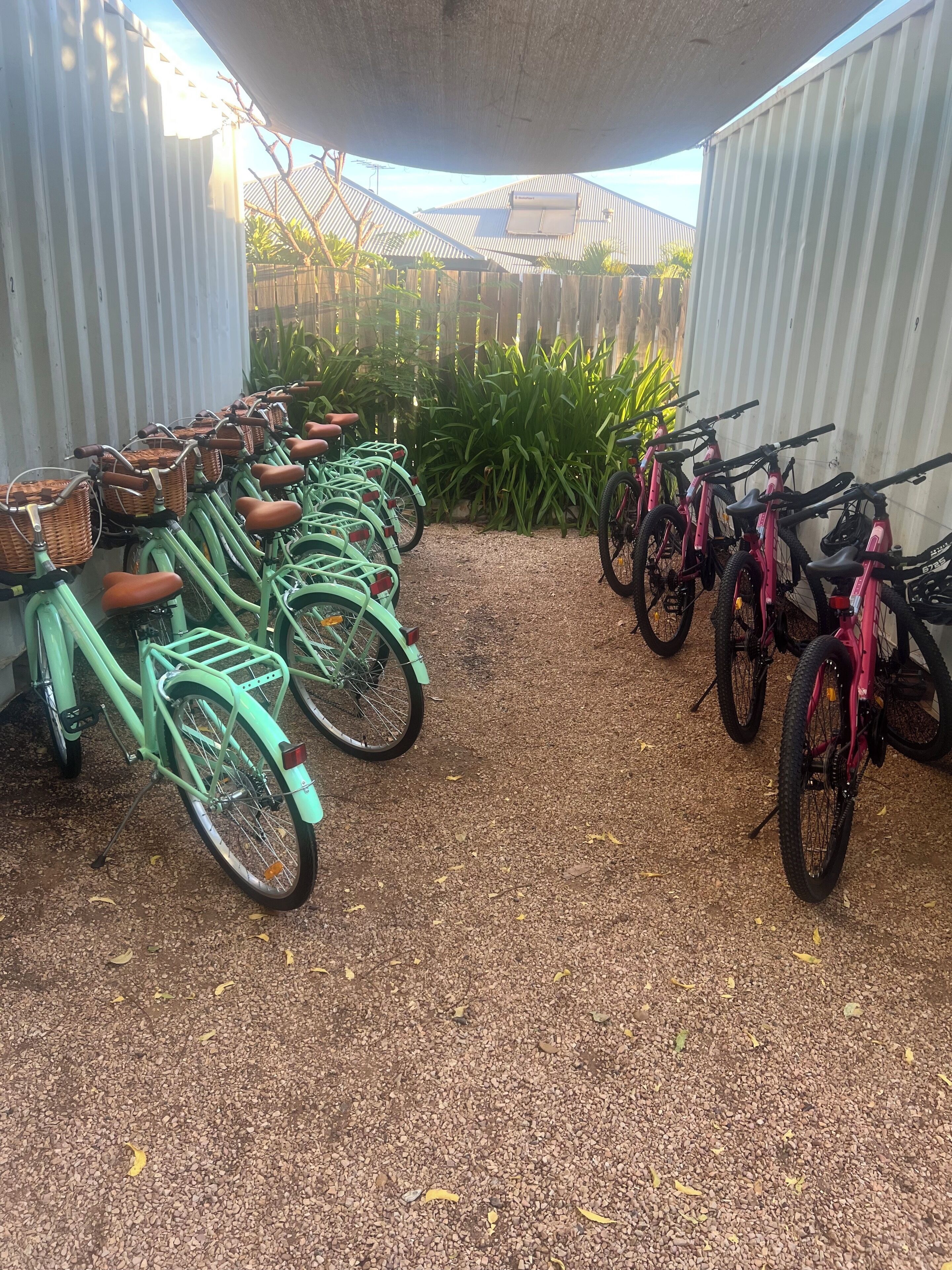 A row of bicycles parked on gravel, with a fence and greenery in the background.