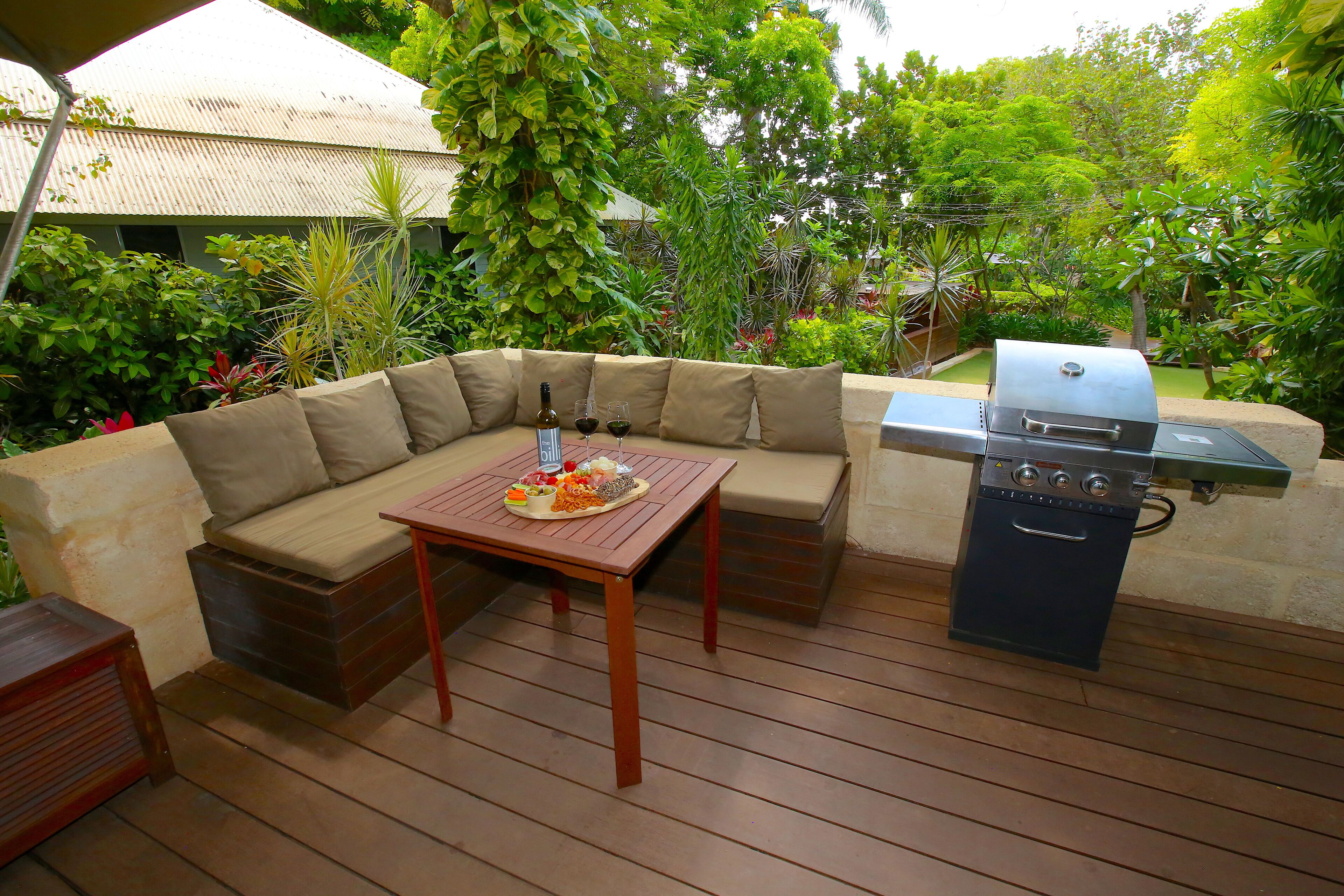 An outdoor patio with a wooden table, a barbecue grill, and a cushioned seating area surrounded by lush greenery.