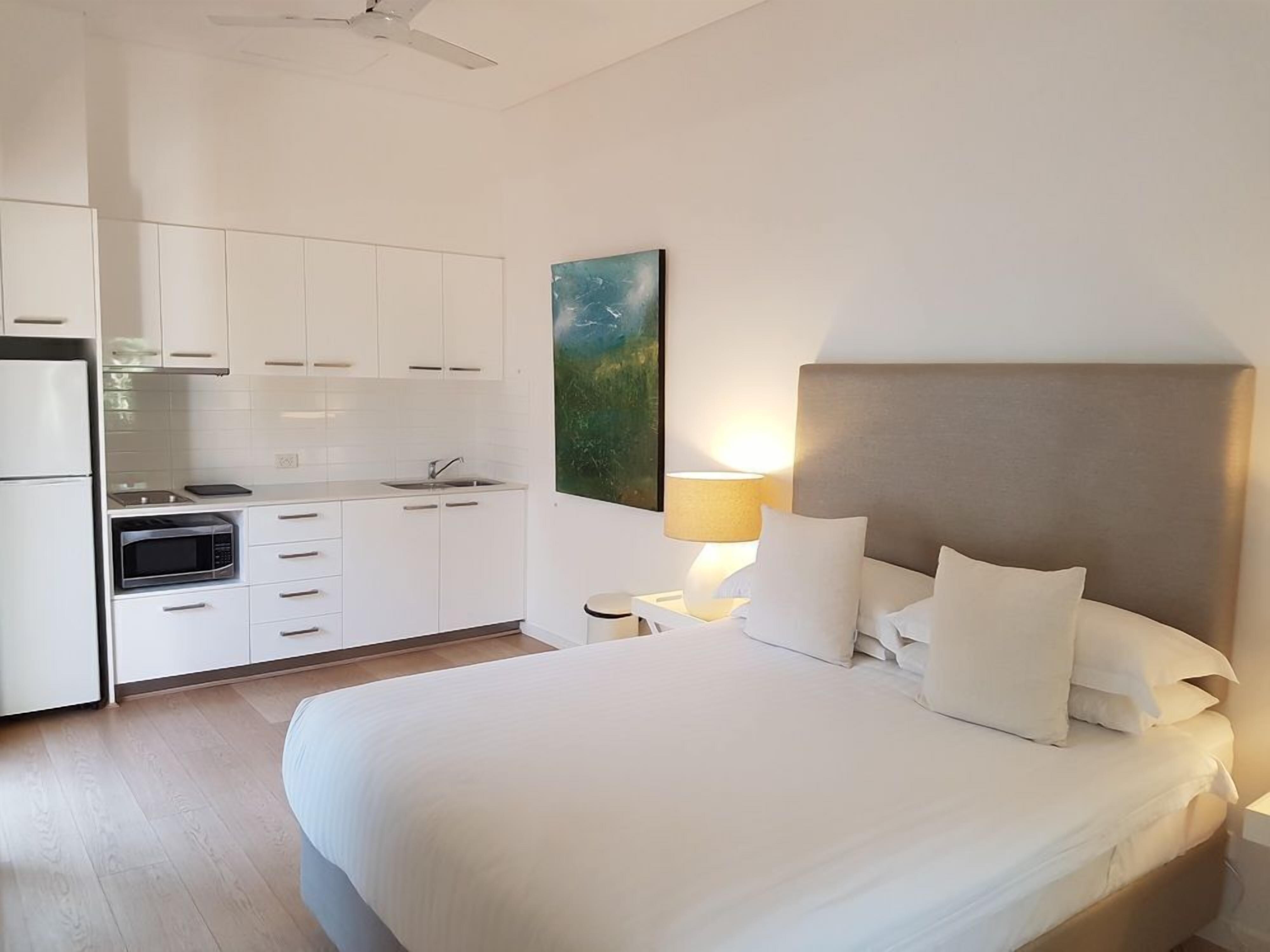 A hotel room with a bed, a nightstand, a lamp, a painting, and a kitchenette.