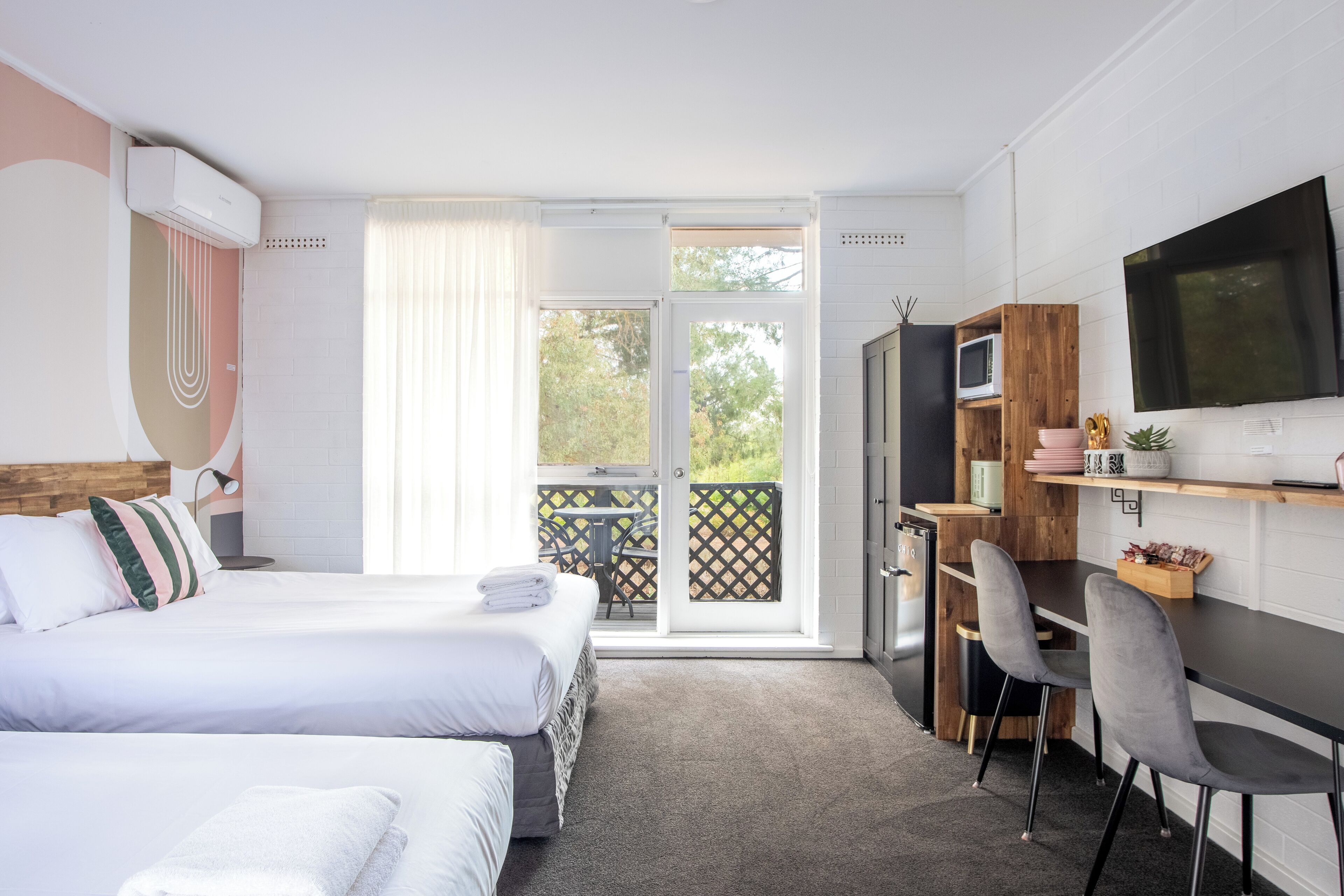 Una habitación de hotel moderna con dos camas, un escritorio, televisión y balcón con vistas a árboles.