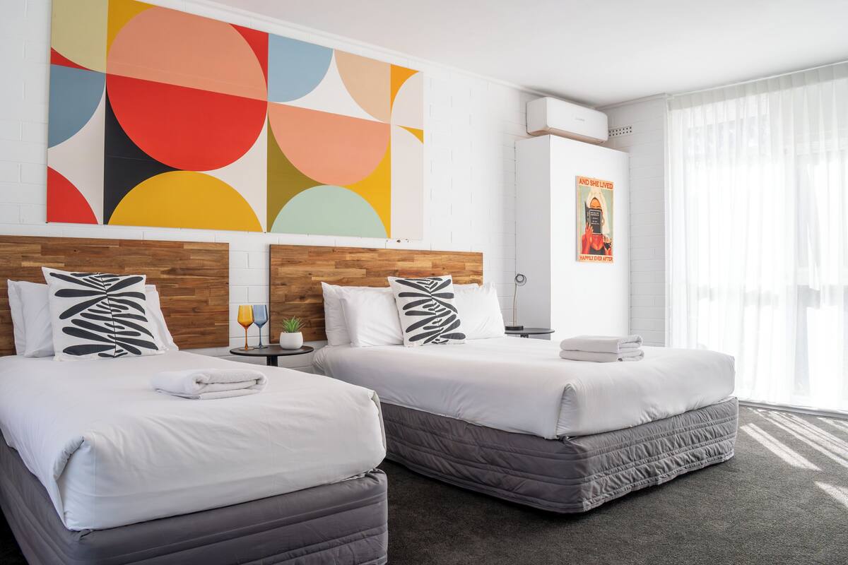 A hotel room with two beds, a colorful abstract painting, a poster, and a window with sheer curtains.