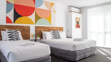 Une chambre d’hôtel avec deux lits, un tableau abstrait aux couleurs vives, une affiche et une fenêtre ornée de rideaux légers.