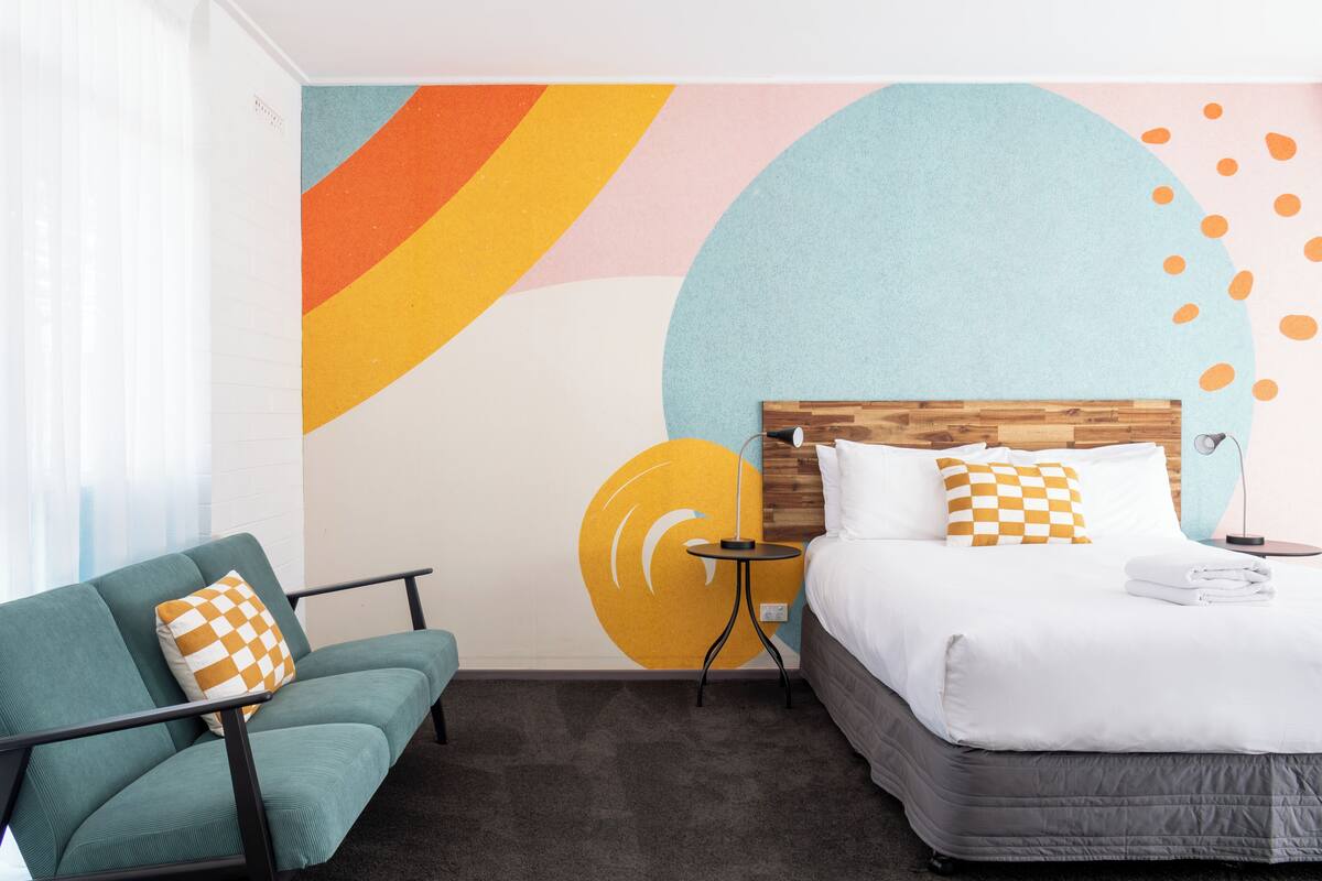 A hotel room with a bed, a sofa, a small table, and a wall with abstract art.