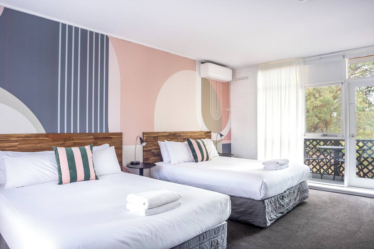 A hotel room with two beds, a wooden headboard, a large abstract wall design, a window with curtains, and a balcony with a table and chairs.
