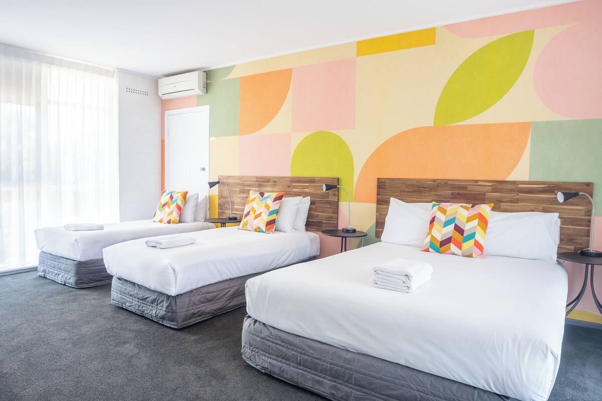 A hotel room with two beds, each with a wooden headboard and white bedding. There is a window with sheer curtains, a wall air conditioner, and a colorful abstract wall design.