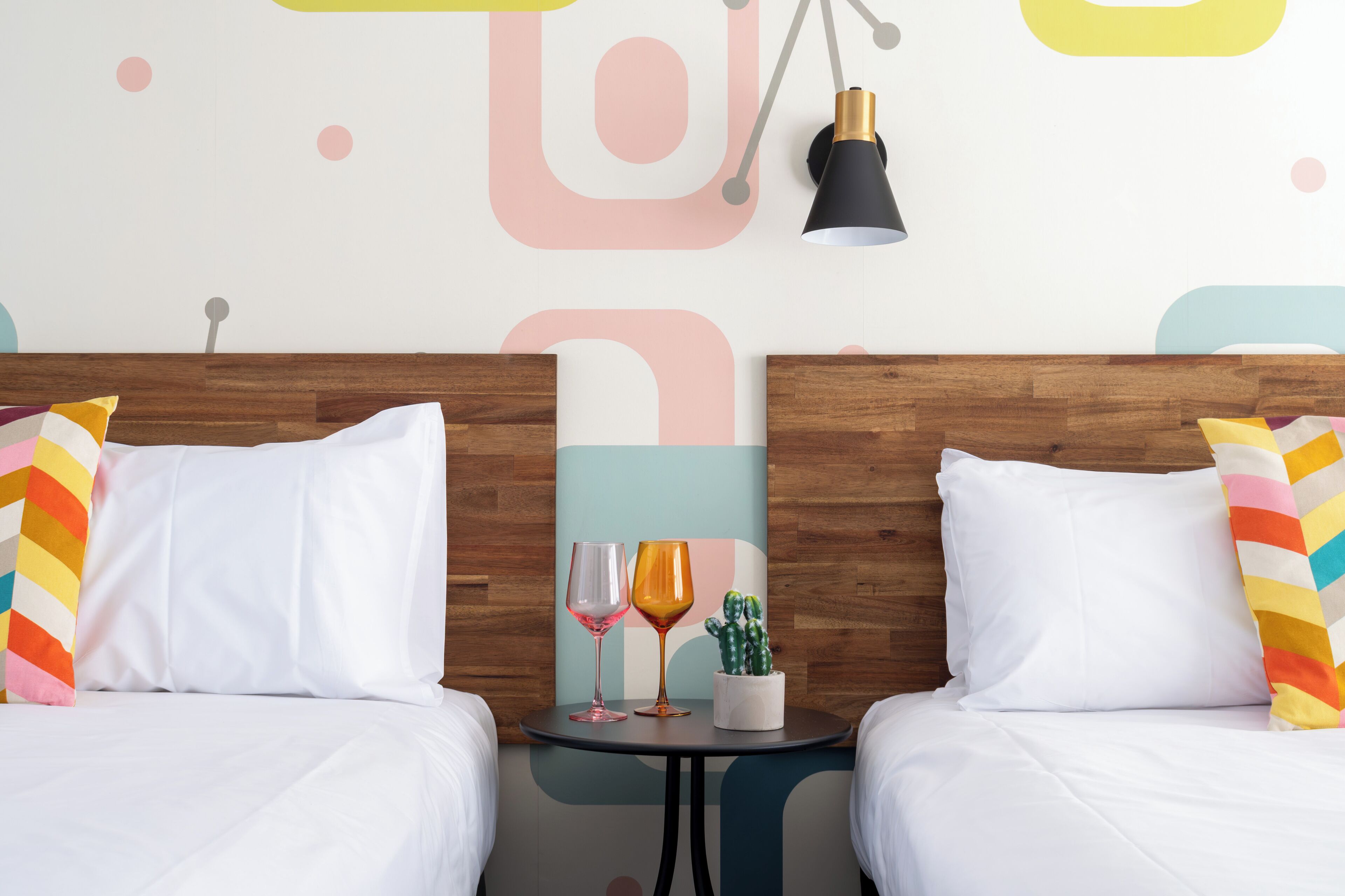 A hotel room with two beds, white bedding, colorful pillows, a small table with two wine glasses and a cactus plant.