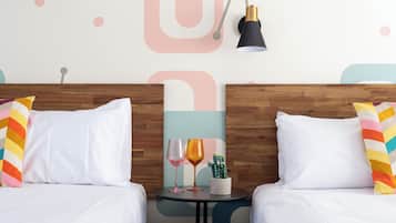 A hotel room with two beds, white bedding, colorful pillows, a small table with two wine glasses and a cactus plant.
