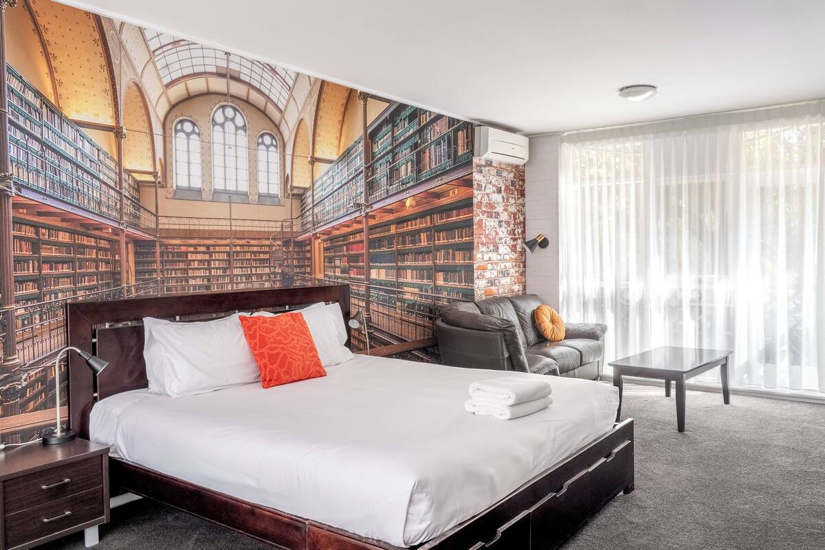 A hotel room with a large bed, a desk, a chair, and a bookshelf mural on the wall.