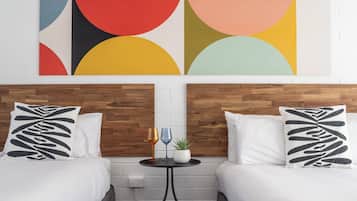 Een hotelkamer met twee bedden, een klein tafeltje met twee wijnglazen en een plant, en een kleurrijk abstract schilderij aan de muur.