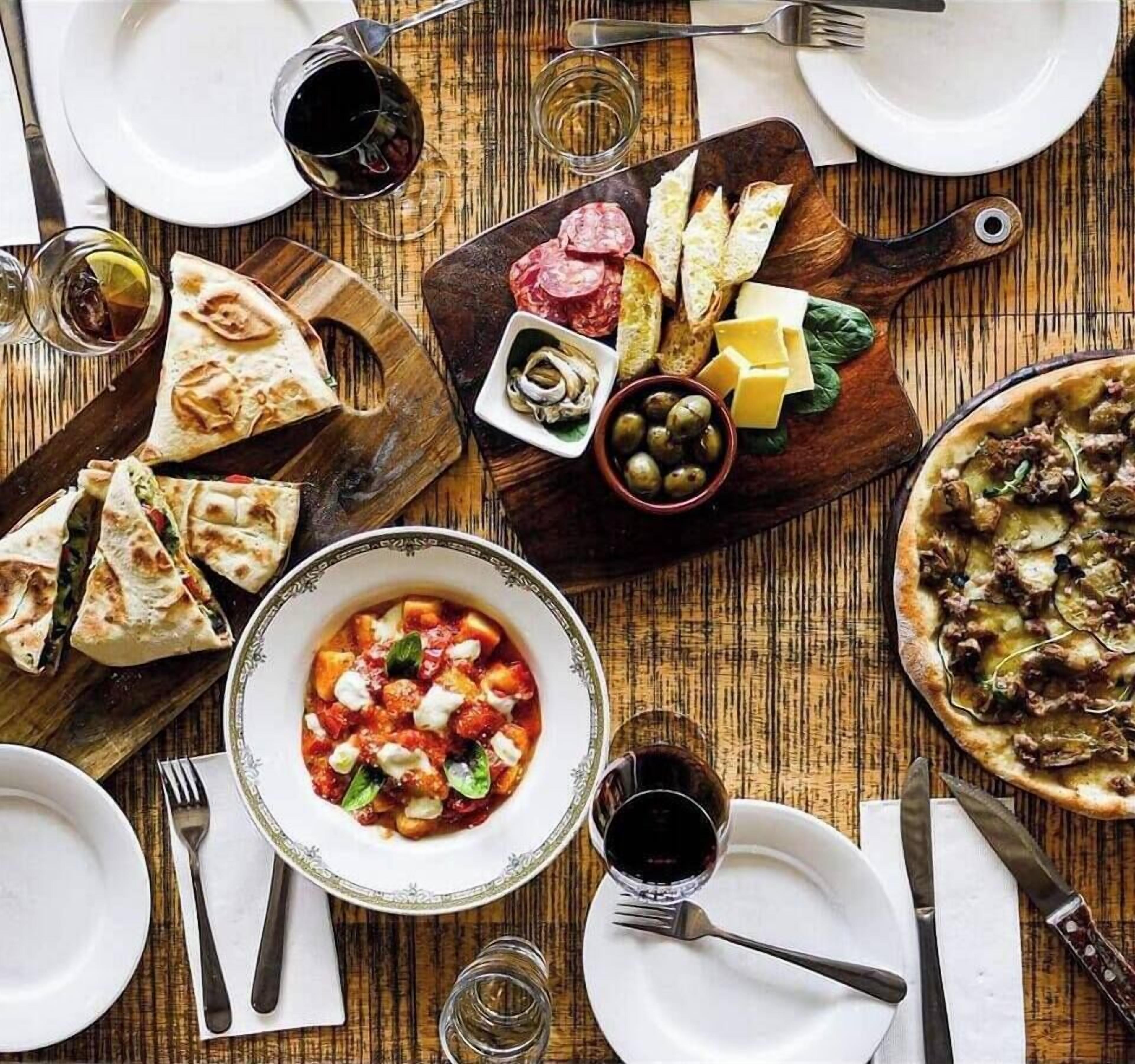 Ein Tisch ist reichhaltig mit Speisen gedeckt, darunter eine Suppe, Pizza sowie eine Auswahl an Käse und Wurstwaren.