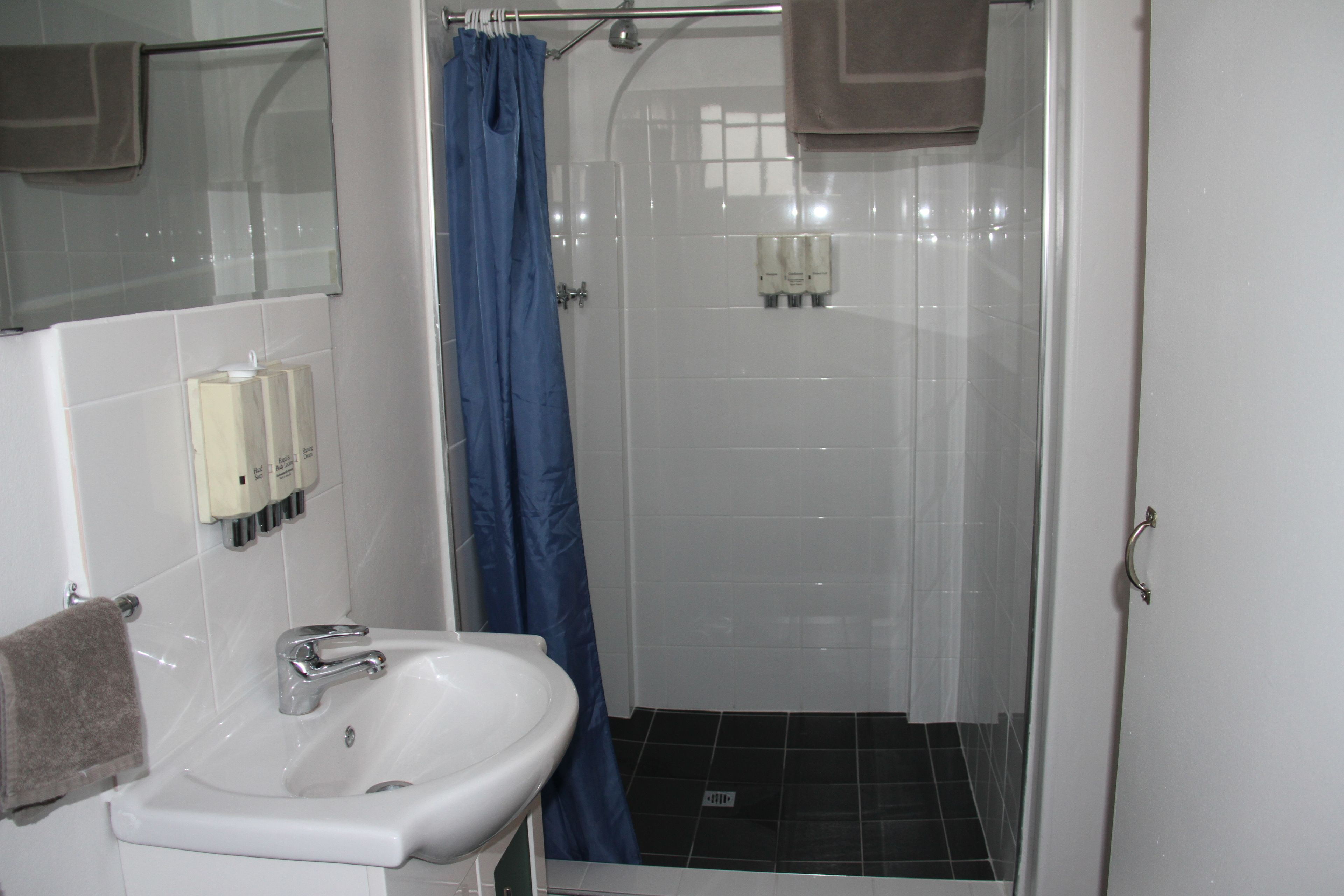 Un baño con un lavamanos blanco, un espejo, una ducha con cortina azul y una puerta blanca.