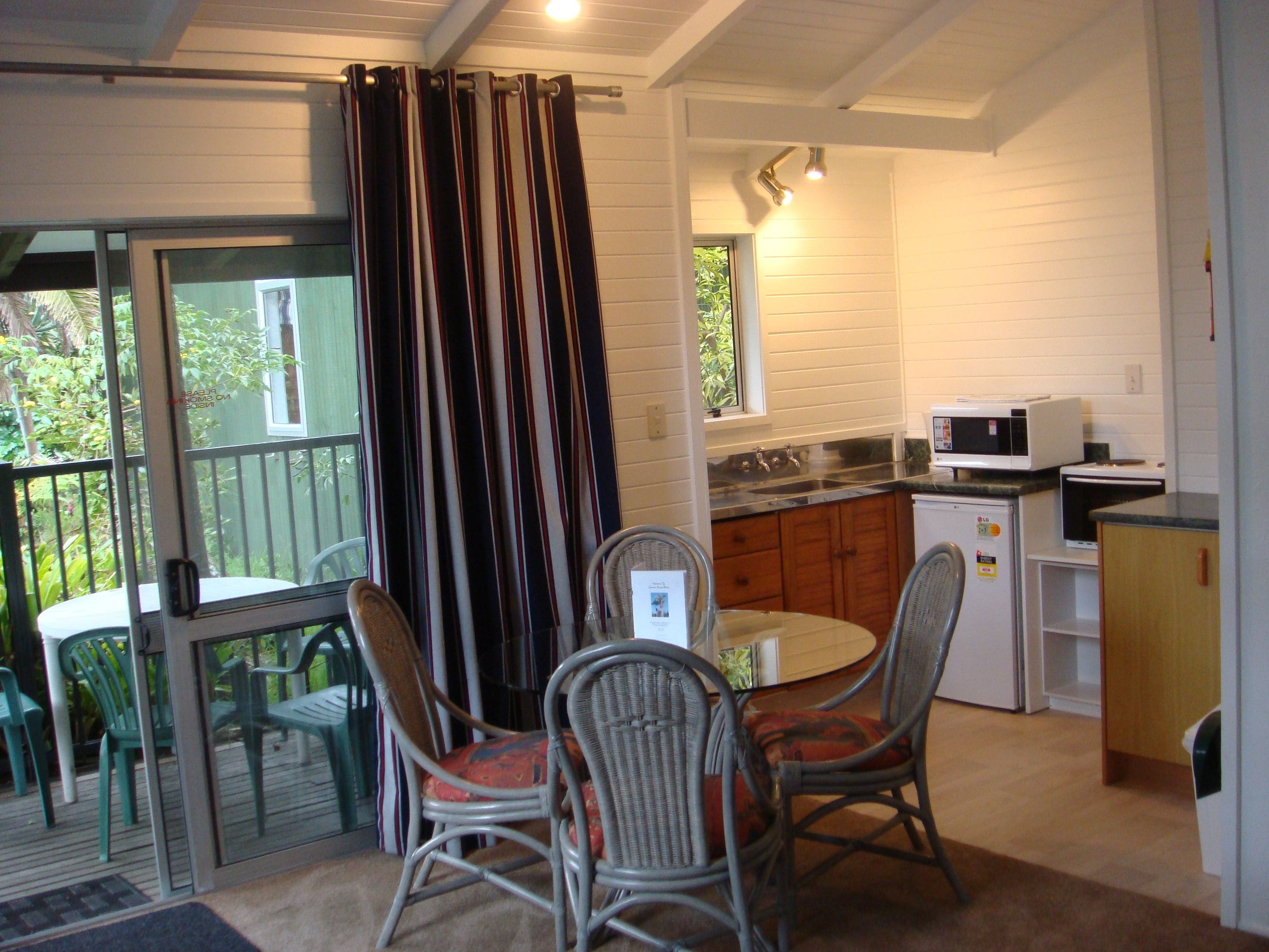 Een keuken met witte kastjes, een magnetron en een spoelbak. Er is een eethoek met rieten stoelen en een glazen tafel.