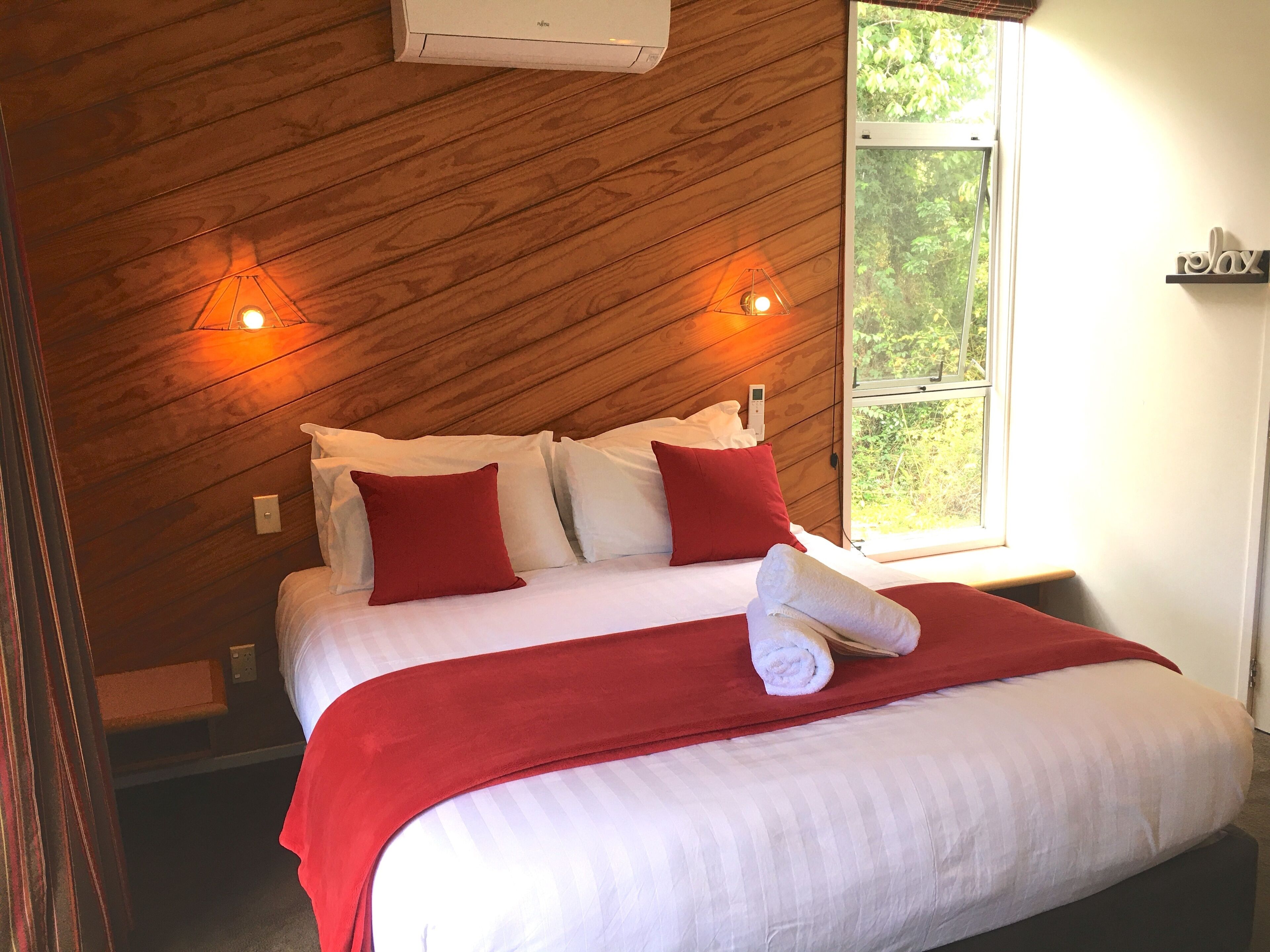 A neatly made bed with white and red bedding, two wall-mounted lights, and a window with a view of greenery.