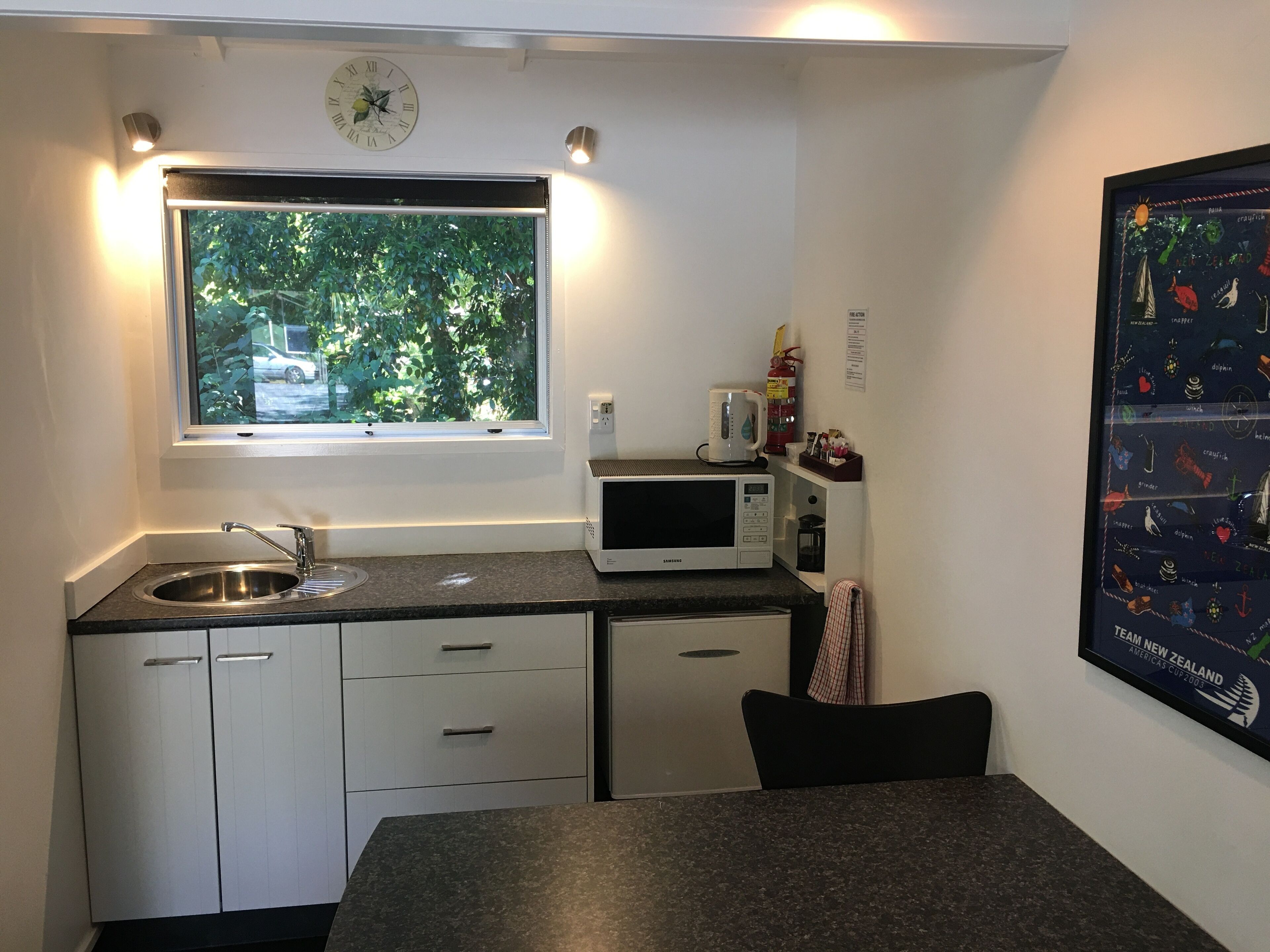 A compact kitchen with a window, a microwave, a sink, and a clock on the wall.