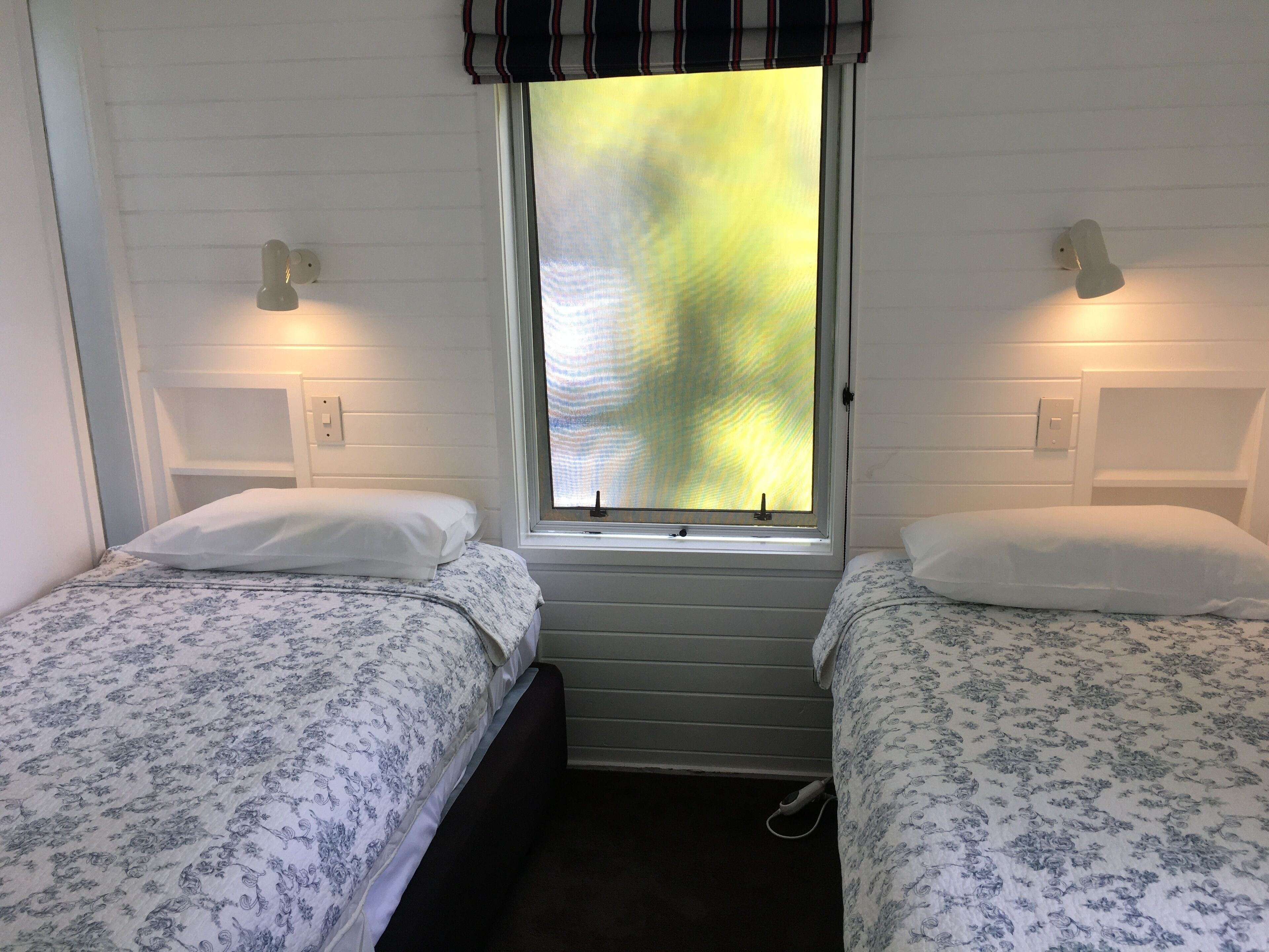 A room with two beds, each with a white pillow and a floral-patterned duvet. There is a window with a view of a yellow-green landscape.