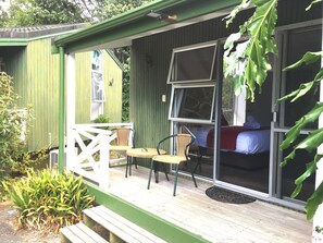 Una estructura pequeña con techo verde, un porche, una cama con manta roja y una ventana con mosquitero.